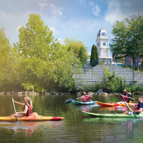 A group of people kayaking on a calm White River, all wearing life vests and enjoying a sunny day. Lush green trees line the riverbank, and a white clock tower rises in the background against a blue sky, creating a scenic and active outdoor setting.