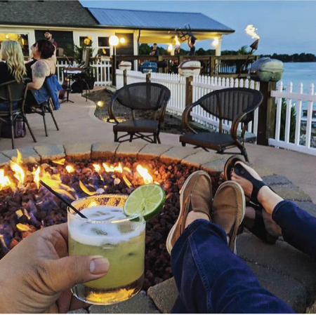 A cozy evening scene at a lakeside restaurant with people relaxing around a fire pit. In the foreground, two people sit with their feet up by the fire, one holding a cocktail garnished with a lime. The background shows other patrons dining on a patio under soft lighting, with views of the lake and tiki torches glowing at dusk.
