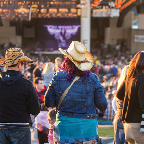 A crowd gathers at an outdoor concert venue during golden hour. In the foreground, two people wearing cowboy hats and denim jackets stand facing the stage, which is lit up with purple lights and a graphic display. The venue is filled with people enjoying the lively atmosphere, with warm sunlight casting a glow over the scene.