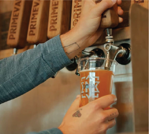 A lively evening scene at a Greek festival with a large crowd gathered under white tents. People are eating, drinking, and socializing in front of a beautifully lit Byzantine-style church with domes and crosses. The sky is dusky blue, and the warm glow from the church windows adds to the festive atmosphere.