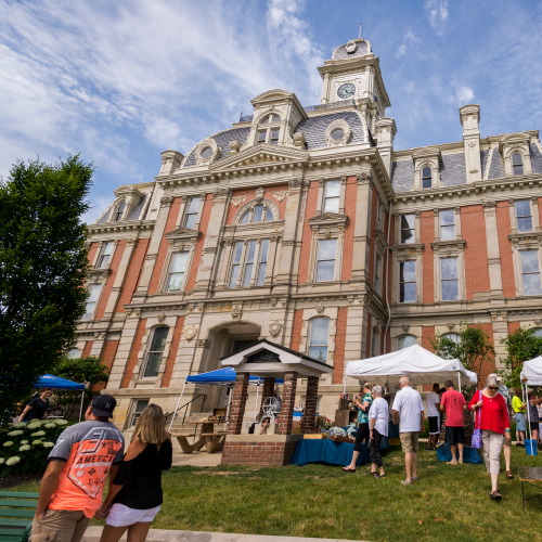 People gather on the lawn of a historic red-brick courthouse with ornate architectural details and a clock tower during a sunny outdoor festival. White tents are set up with vendors and tables, and attendees stroll through the event, enjoying the festive atmosphere. The sky is partly cloudy, adding to the charm of the summer day.